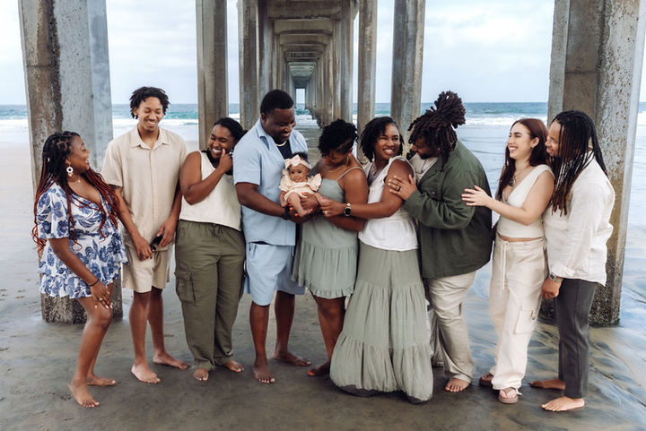 Family under a beach pier smile around a baby, with ocean and concrete pillars behind them.