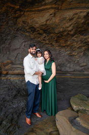 Family of three in front of a rocky cave. Man in white shirt holds child in white; woman in green dress is pregnant. All smiling warmly.