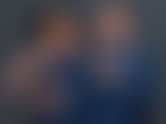 Couple in matching navy outfits smiling warmly at each other against a dark backdrop. The woman has her hand on the man's shoulder.