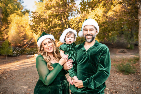 Family in green velvet outfits and Santa hats smiles in a forest. Sunny autumn background with yellow leaves creates a warm, festive mood.