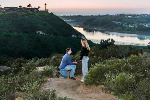 Romantic sunset proposal in San Diego as man kneels with ring box overlooking coastal lagoon