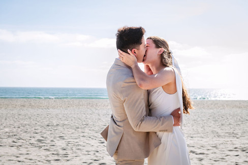 Close-up of a bride and groom sharing a romantic first kiss during their Coronado Beach wedding photoshoot in San Diego, California, with the ocean and sandy beach in the background.