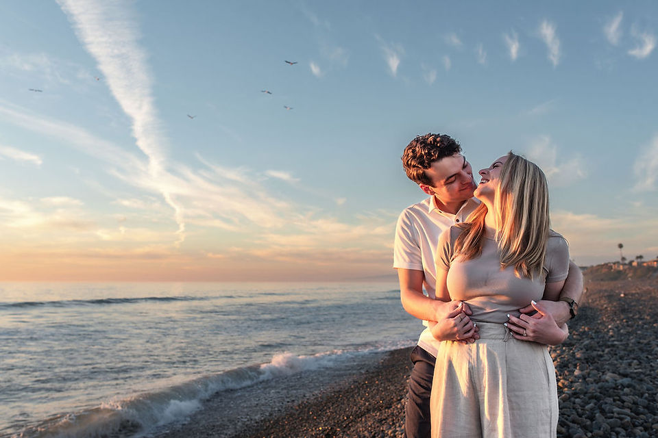 Couple standing on beach shoreline, man hugging woman from behind looking out at ocean