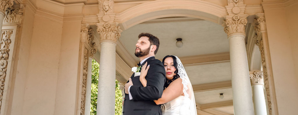 Bride in white lace wedding gown with veil hugging groom in black suit under ornate Balboa Park columns.