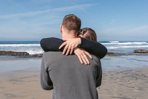Engaged woman hugs fiancé from behind displaying ring on sunny beach in Cardiff-by-the-Sea