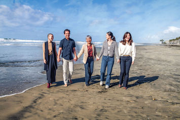 Family walking hand in hand on a sunny beach, smiling. They wear casual clothes. The ocean and blue sky are in the background.