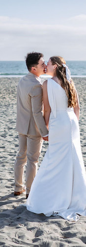 Back view photograph of the newlywed couple kissing in Coronado beach. They are holding hands and facing the ocean, with the bride and groom seen from behind as they kiss. Their figures cast distinct shadows on the sand. The background is a calm ocean and horizon with faint clouds.
