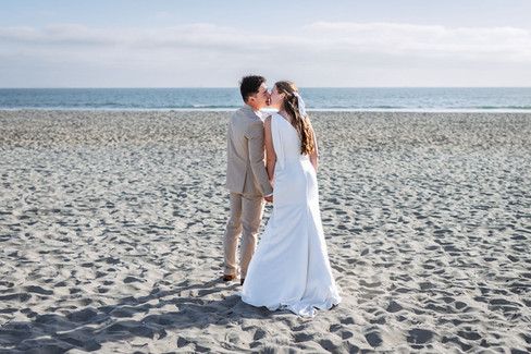Full-length shot of a bride and groom kissing on the sand during a Coronado Beach wedding photoshoot in San Diego, California, with a wide view of the ocean and bright blue sky.