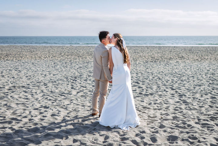Back view photograph of the newlywed couple kissing on Coronado beach. They are holding hands and facing the ocean, with the bride and groom seen from behind as they kiss. Their figures cast distinct shadows on the sand. The background is a calm ocean and horizon with faint clouds.