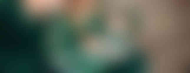 A couple and a baby, all in green velvet outfits, sit on a blanket outdoors. The baby smiles at the woman, surrounded by fallen leaves.