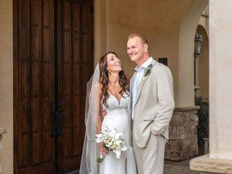 Bride in white gown and groom in beige suit smiling joyfully outside by wooden doors, bride holding white flowers. Neutral-toned setting.