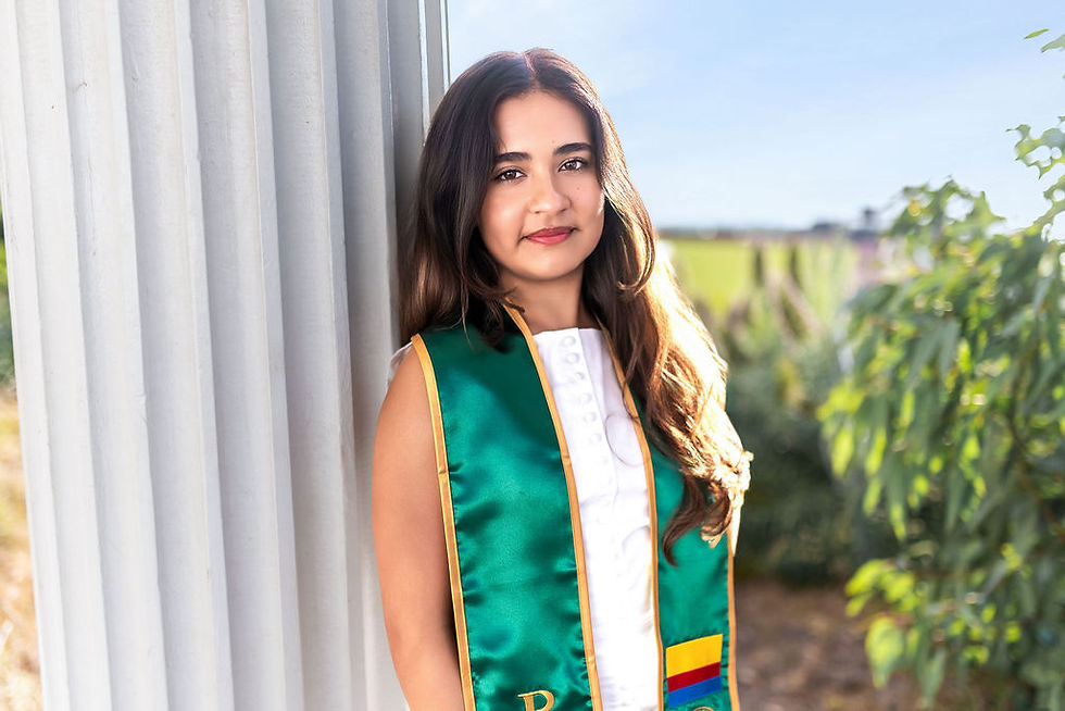 Young woman in a green-and-gold sash over a white dress stands by white columns outdoors in bright sunlight.