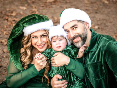 Family wearing green Santa hats and outfits, smiling in a park. The mood is festive and joyful, with brown leaves on the ground.