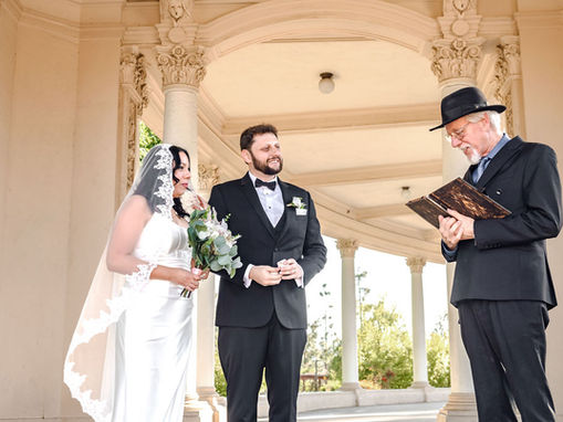 Bride in white wedding gown and lace veil standing with groom in black tuxedo and bow tie while officiant in black suit and hat reads vows under Balboa Park arches.