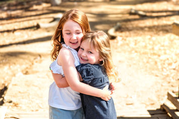 Two young girls hugging and smiling in a sunlit park with a sandy path. One wears a white top, the other dark blue, showing joy and affection.