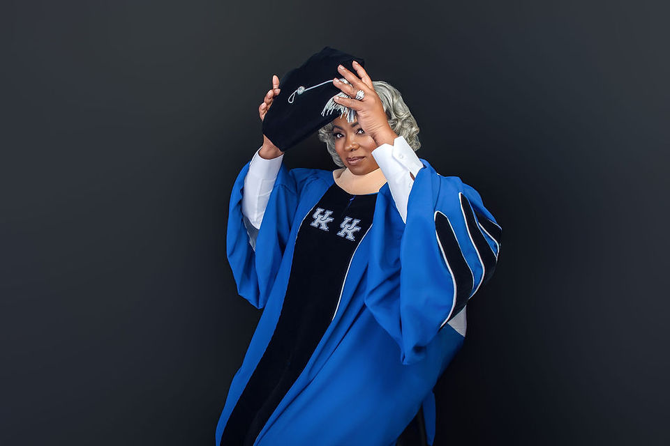 Person in a blue academic robe adjusts cap, against a dark background. The robe has "HK" text, conveying a confident, celebratory mood.