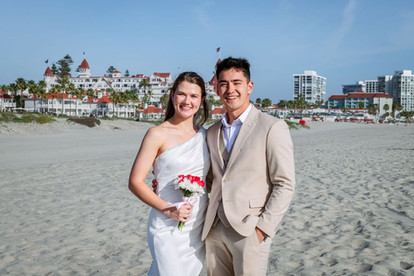 Smiling bride in a white one-shoulder wedding dress and groom in a tan suit posing on Coronado Beach, San Diego, with the Hotel del Coronado in the background.
