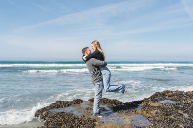 Joyful fiancé lifts woman in the air celebrating engagement on tide pools at Cardiff State Beach San Diego