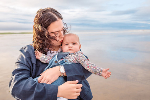 Woman holds a baby at a beach, kissing the baby's head. The baby wears floral-patterned clothes and a denim outfit. Calm, serene mood.