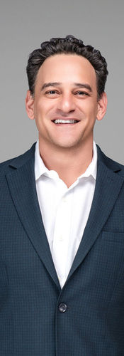 Man with short curly dark hair wearing dark gray blazer over white dress shirt, standing against gray background, smiling.