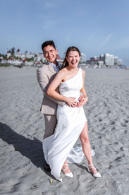 Playful vertical photograph of a groom holding his laughing bride from behind, posing on Coronado Beach with Hotel del Coronado visible in the background.