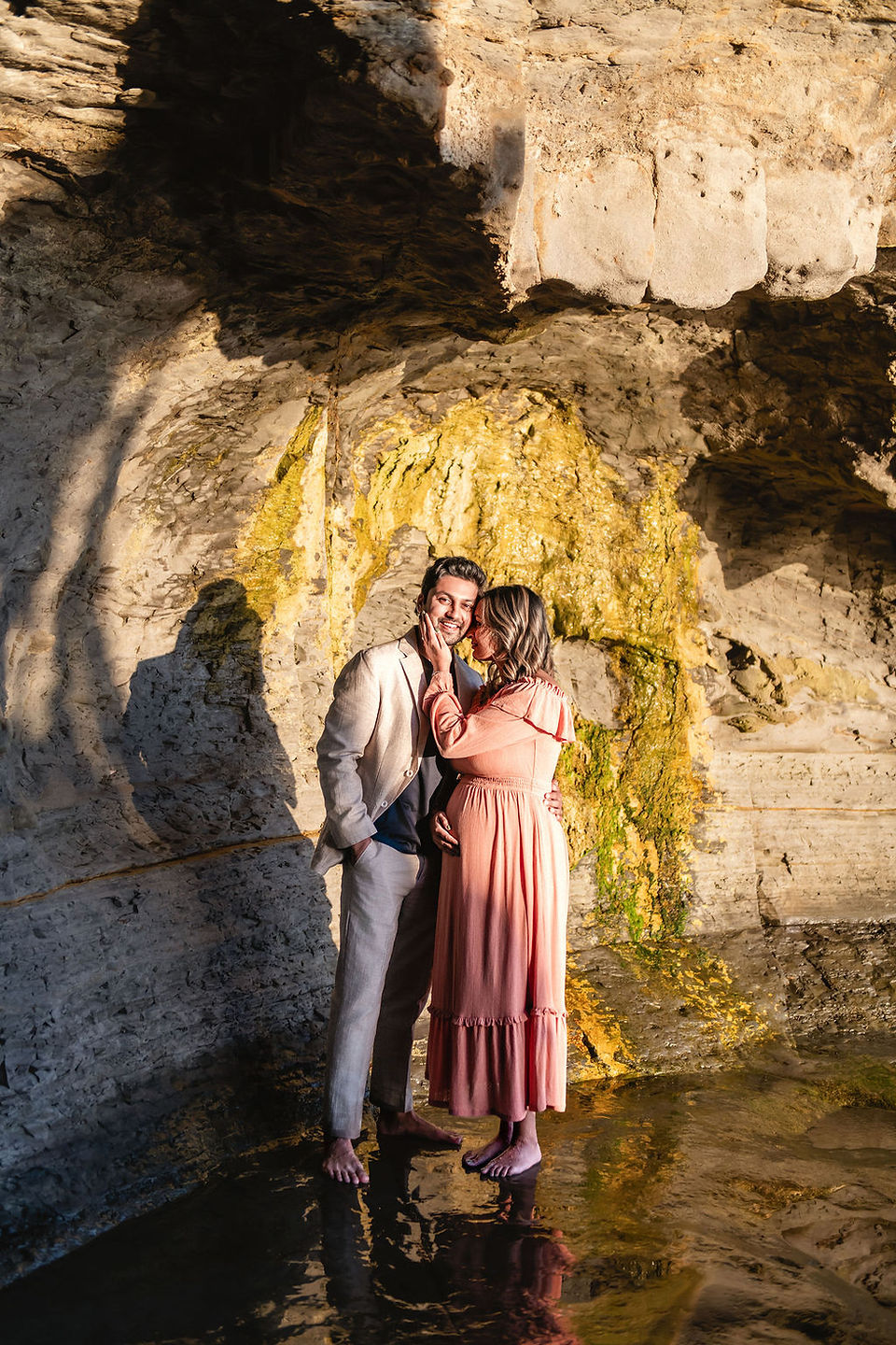 Couple embraces against a rocky, golden-lit cliff. Woman in peach dress; man in beige suit. Barefoot, relaxed mood, reflective water below.