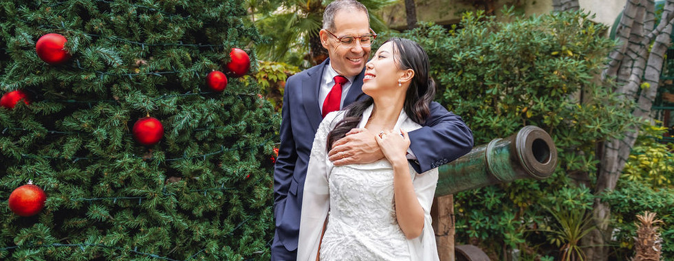A couple embraces joyfully by a decorated Christmas tree with red ornaments; set in a lush garden with a historic cannon nearby.