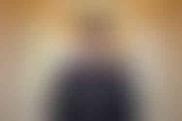 Bride’s hands on groom’s shoulders from behind under Balboa Park columns, groom in black suit, bride in white lace wedding gown.