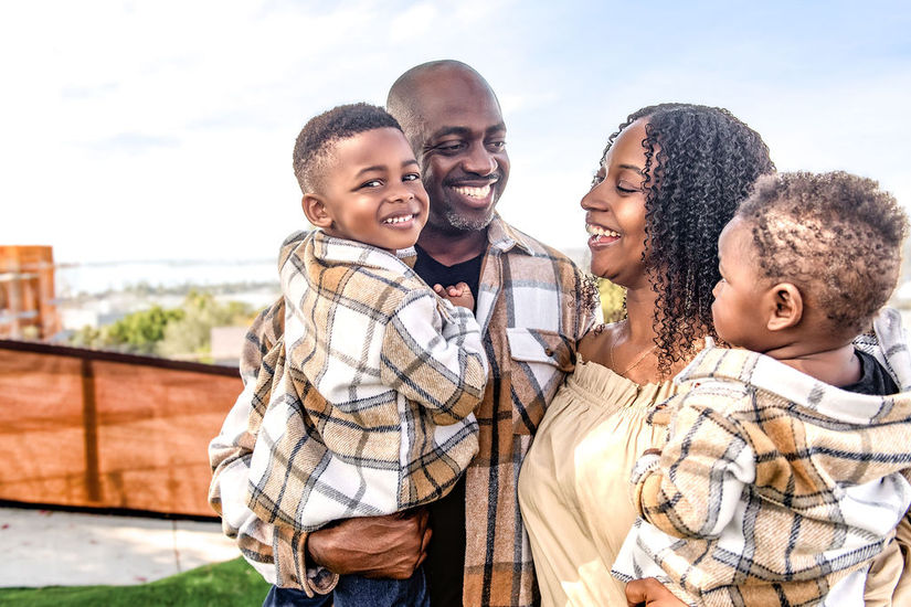 Family smiling outdoors, parents holding two children. Background shows a bright, sunny landscape with trees and a clear sky.