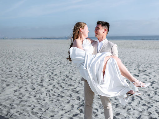 Wide-angle, vertical photograph of the groom carrying the bride in his arms across a wide, sandy Coronado beach. The groom is wearing a beige suit and brown shoes, and the bride is in her white wedding dress, holding the fabric. They are looking at each other and smiling. The horizon of the ocean and a blue sky with faint clouds are behind them.
