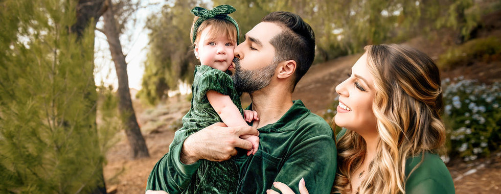 Family in green velvet outfits and hats, kissing a child outdoors in a park with autumn leaves. Warm, joyful atmosphere.