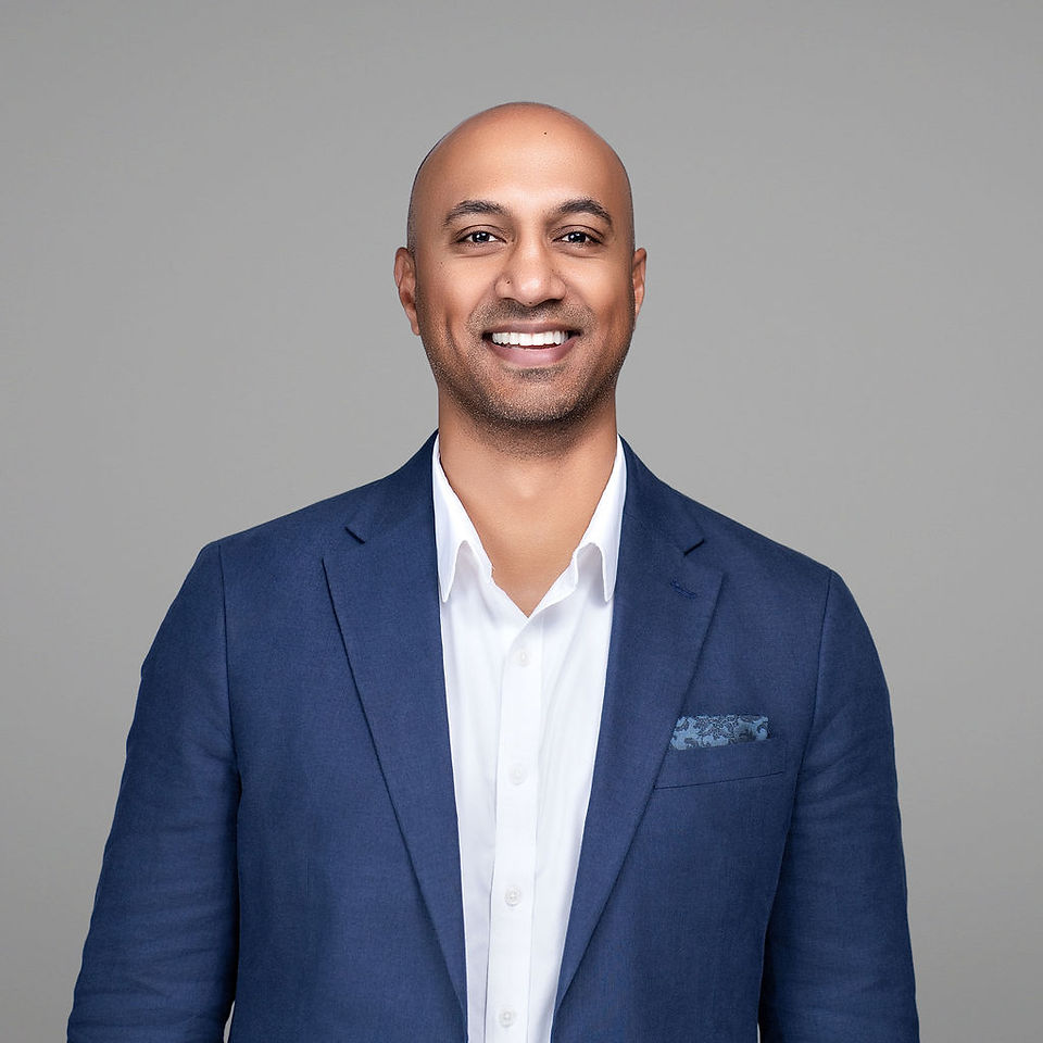 Man wearing navy blue blazer over white dress shirt with pocket square, standing against gray background, smiling.