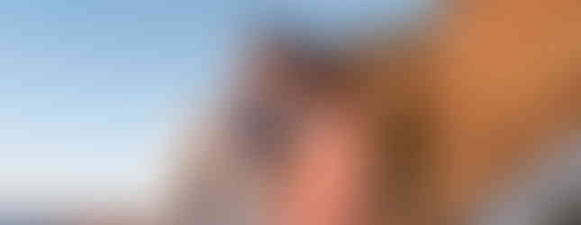 A couple stands on a seaside railing with rocky cliffs and blue sky. Woman in a peach dress, man in a beige jacket, smiling warmly.