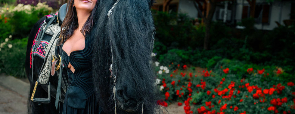 Confident woman in black romper embracing a black Friesian horse beside red flower garden at a Murrieta ranch birthday session