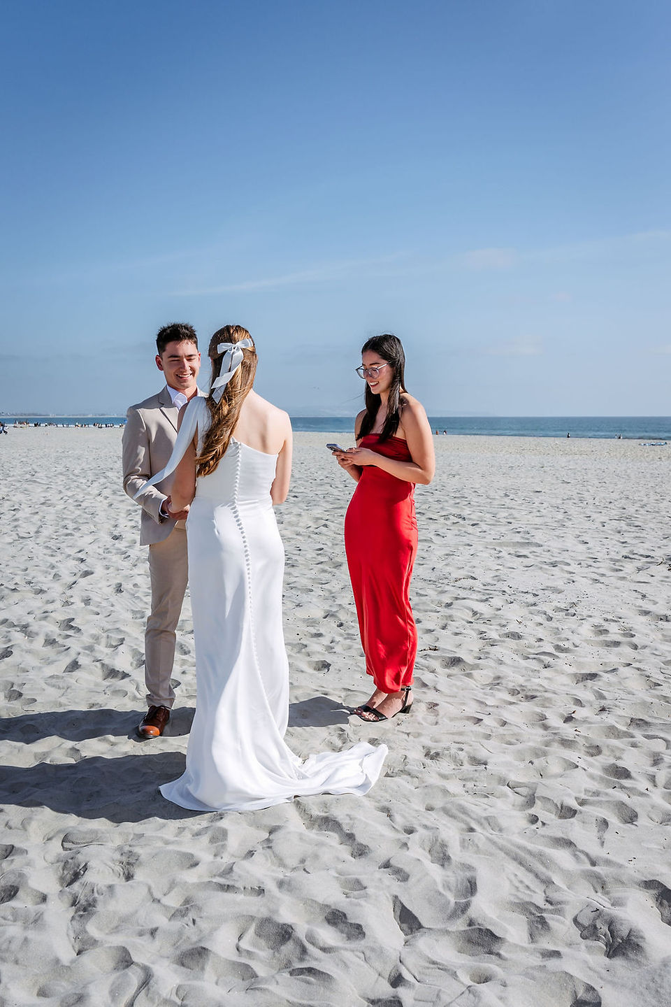 Bride and groom holding hands during an intimate Coronado Beach wedding ceremony in San Diego while an officiant in a red dress reads from a phone on the sand.