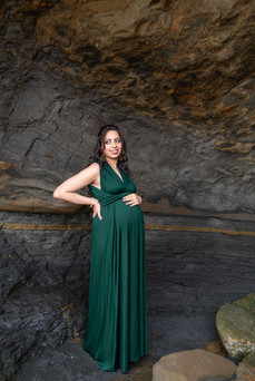 Pregnant woman in a green dress cradles belly, smiling, in a rocky cave setting. Earthy tones in background, serene mood.