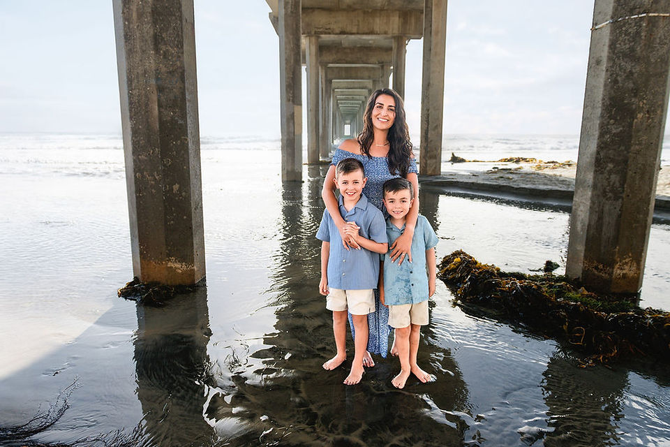 Mother with two young sons in blue outfits standing under Scripps Pier during a La Jolla family photography session