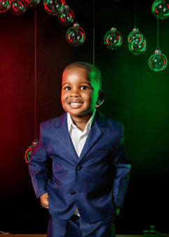 Boy in blue suit smiles against a black background with red and green lighting and hanging glass bubbles.