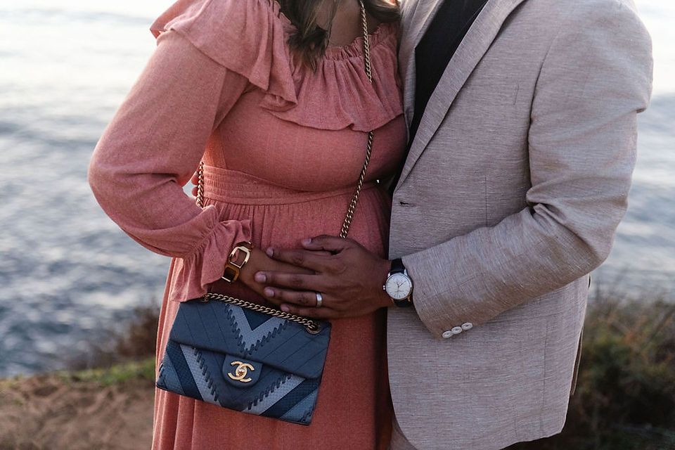 Couple embracing by the water, woman in pink dress with a designer purse, man's hand on her belly, suggesting a tender moment.