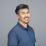 Man with short dark hair with gray streaks and beard wearing navy blue polo shirt, standing against gray background, smiling.