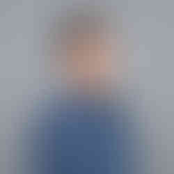 Man with short dark hair with gray streaks and beard wearing navy blue polo shirt, standing against gray background, smiling.