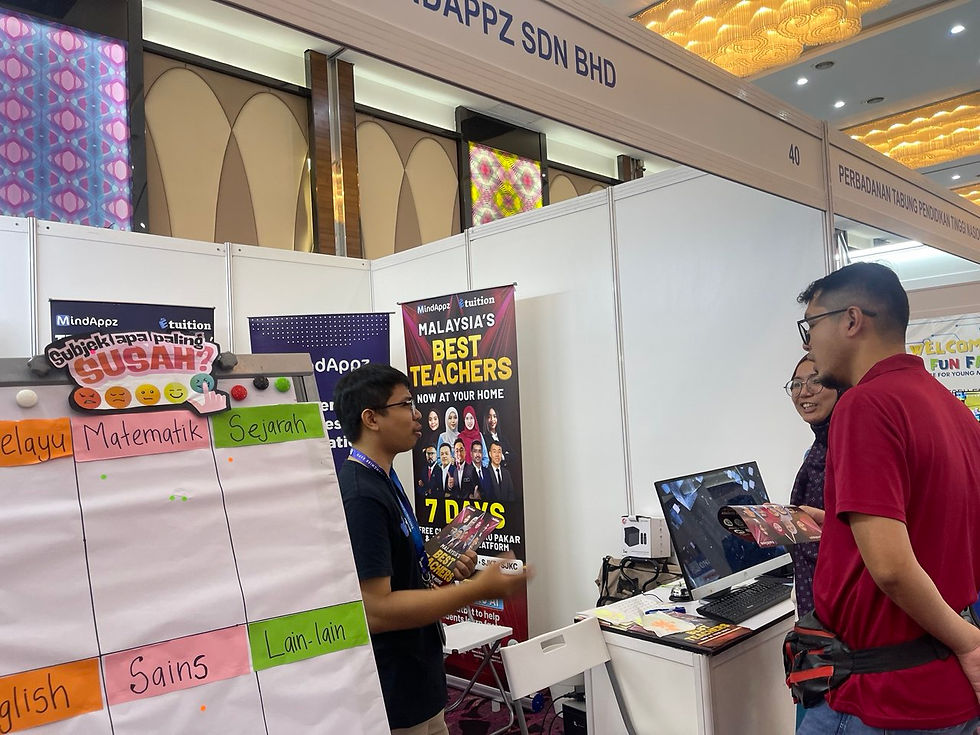 A man at an educational booth talks to two people. A board displays subjects. Bright lights and colorful banners create a lively atmosphere.