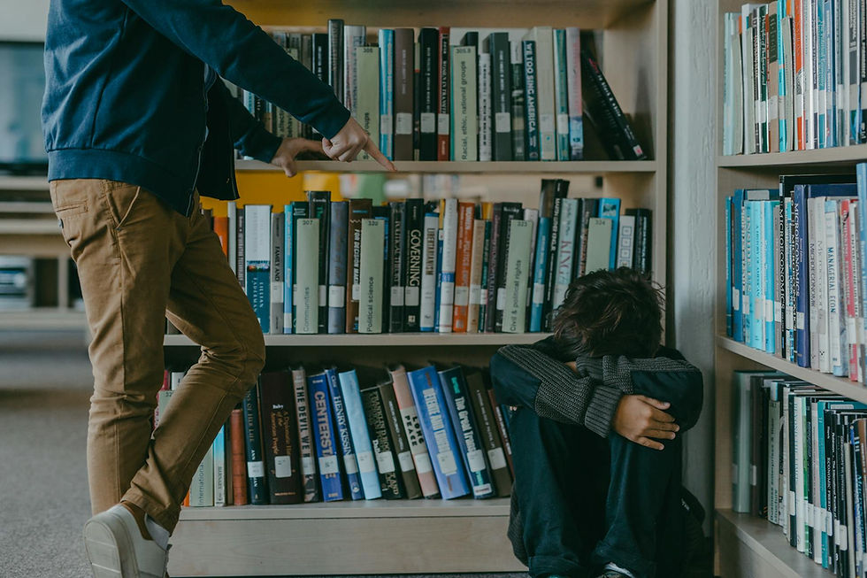 Person pointing at a seated individual in distress, surrounded by bookshelves. The setting is a library with colorful book spines visible. Mood: tense.