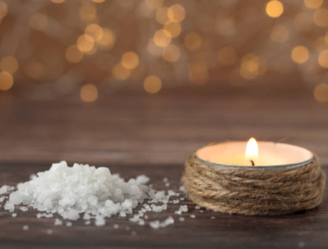 A lit candle wrapped in twine sits on a wooden surface beside a small pile of coarse salt, with soft golden lights blurred in the background.