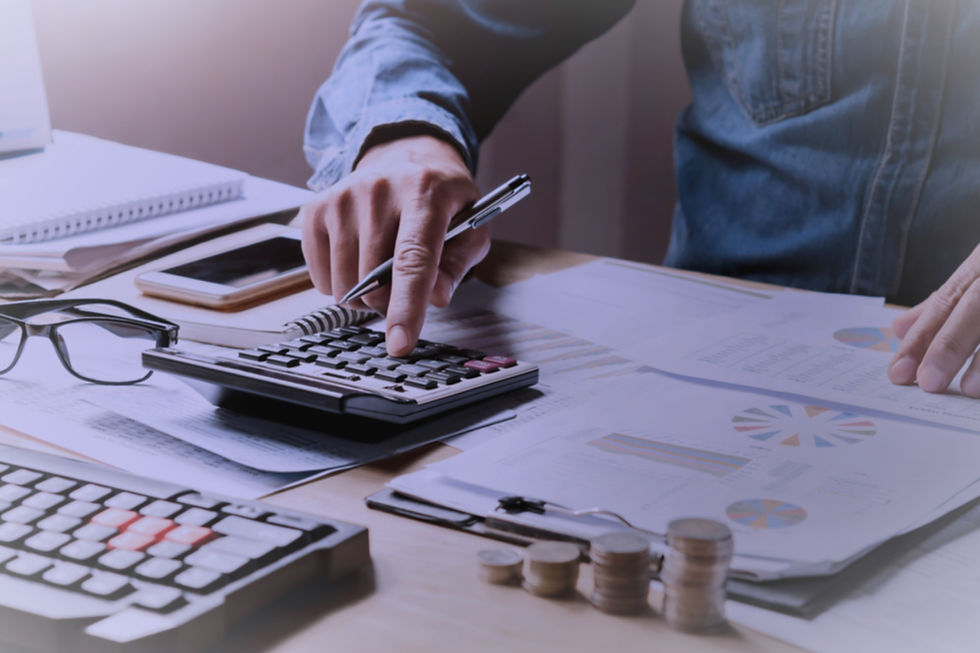 A man in a blue shirt surrounded by paper, coins and electronics is using a calculator