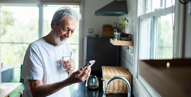 Senior man smiling at smartphone while drinking water at home