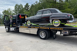 Black vintage car secured on a flatbed tow truck from JJ's Road Towing & Recovery, parked on a road with trees in the background.