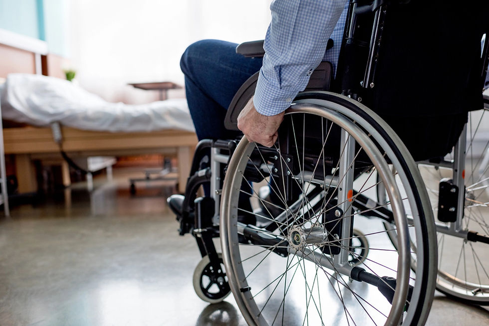 Photograph showing a person seated in a wheelchair gripping the wheel, positioned in a room with a bed and wooden furniture in the background. The scene highlights mobility assistance and accessibility within a long-term care setting