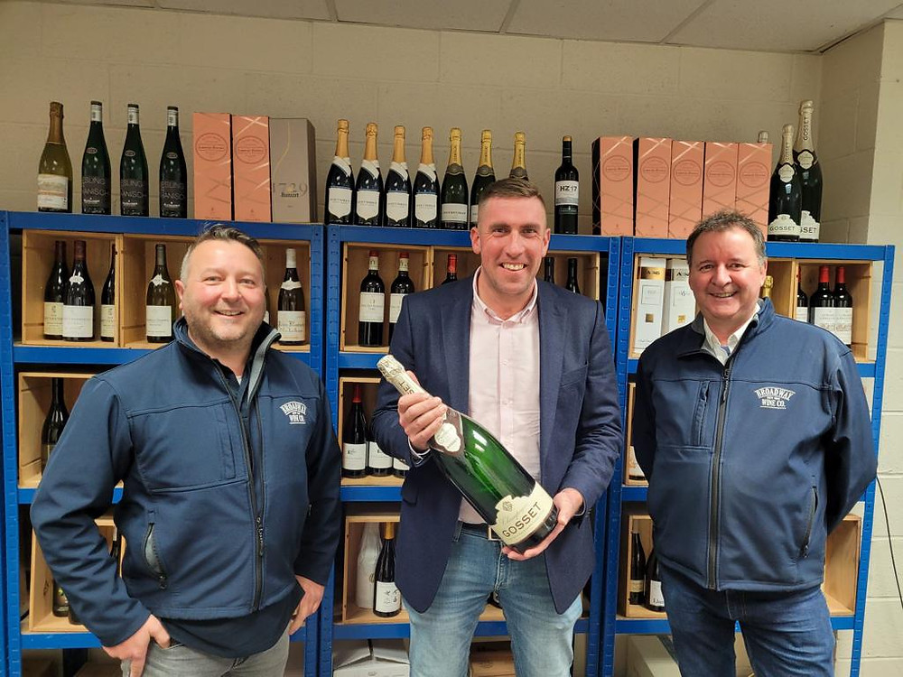 Three men, smiling. The one in the middle is holding a large bottle of champagne. Behind them are shelves full of bottles of wine.
