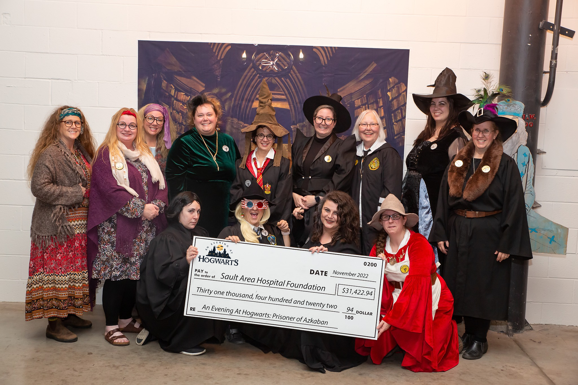 A group of 13 volunteers from An Evening at Hogwarts pose dressed as characters from the Harry Potter universe. They are gathered around a large cheque in the amount of $31,422.94 to SAH Foundation.